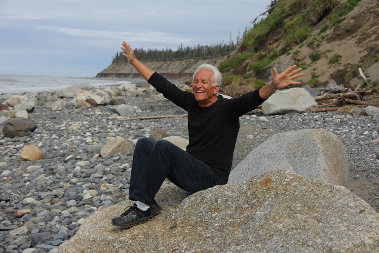 Happy older man sitting on a rock on the beach, arms spread out, smiling.