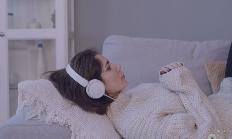 Woman relaxing on sofa wearing headphones and listening to online hypnotherapy.