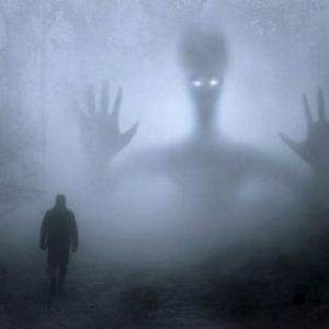 fantasy-2847724_1920 Dark misty wood with shadowy outlines of a man's silhouette walking and a larger, strange looking figure in the background, with piercing white eyes.
